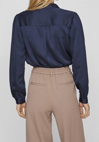 Camicia lunga in satin blu navy con pletature posteriori, abbinata a pantaloni beige a vita alta e gamba larga con tasche posteriori e vita elastica.