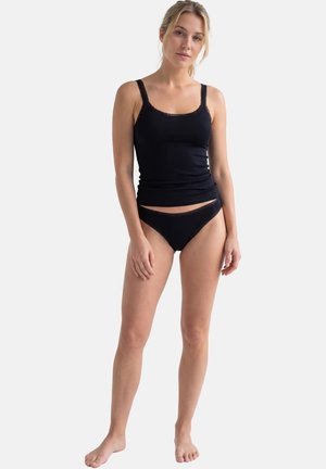 Frau steht barfuß und trägt ein schwarzes, mit Spitze besetztes Negligé und passende Unterwäsche vor einem schlichten weißen Hintergrund.