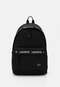 Musta kangasreppu, jossa valkoinen “LACOSTE” -logovyö, etuvetoketjutasku ja brodeerattu krokotiililogo alhaalla oikealla.