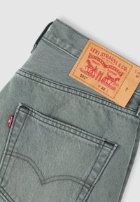 Διπλωμένο γκρι τζιν Levi's 501 που δείχνει την πίσω τσέπη, την κόκκινη ετικέτα Levi's και το ταμπελάκι από δέρμα σε ταμπά χρώμα με το μέγεθος W32 L στη ζώνη της μέσης.