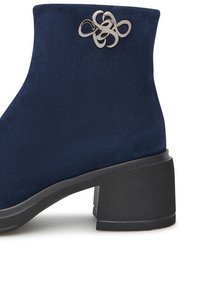 Stivale alla caviglia in suede blu navy con tacco a blocco, caratterizzato da un accento decorativo metallico in un design stilizzato a nodo sul lato.