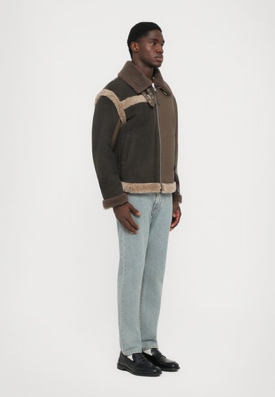 Braune Jacke aus Schaffell mit einem kontrastierenden beigen Panel, Reißverschluss und zwei Seitentaschen, kombiniert mit hellblauen Jeans und schwarzen Slippern.