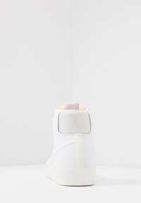 Valkoinen korkea tennari, jossa on teksturoitu takapaneeli, keltaisia korosteita ja Nike-logo kantapäässä. Silkkinen nahkamateriaali ja pehmustettu kaulus.