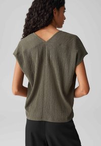 Olive green blouse with a textured, wrinkled finish. Cap sleeves and a wide V-neck at the back, styled with loose-fitting design.