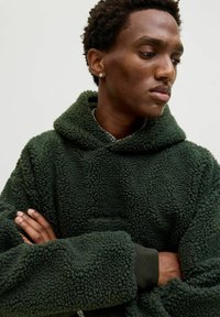 Sweat à capuche en polaire vert foncé avec une surface texturée et une grande capuche, comprenant une poche kangourou fonctionnelle et des poignets côtelés.