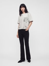 Woman with black wavy hair wears a light gray graphic t-shirt and black straight-leg pants, standing with one hand in pocket on plain white background.