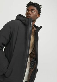 Chaqueta negra impermeable con capucha y un interior marrón en contraste. Cuenta con cierre con marca, tejido texturizado y un diseño elegante.
