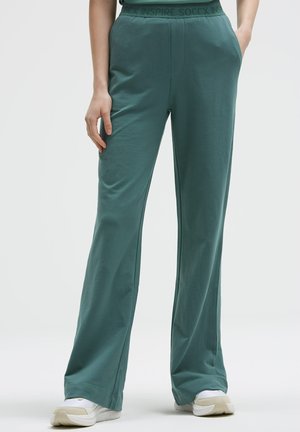 Person trägt lockere türkisfarbene Hose mit elastischem Bund und weißen Sneakers, steht mit einer Hand in der Hosentasche vor einfarbigem Hintergrund.