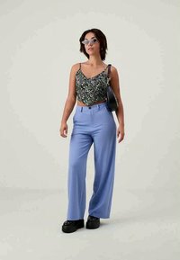 Mulher vestindo um top cropped preto com flores, calças largas azuis claras, sapatos de plataforma pretos, óculos de sol estilo gato, e carregando uma bolsa preta de ombro.