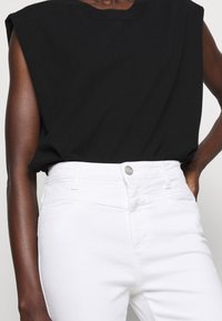Top negro sin mangas con un ajuste relajado y hombros anchos, combinado con pantalones blancos de cintura alta que cuentan con un botón plateado y bolsillos.