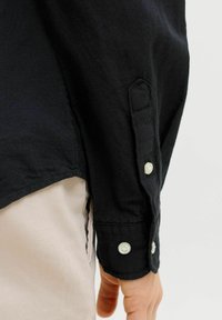 Musta nööpidega särkivars, mis on kantud beežide pükste peale, näidates randme ja käsivarre alumist osa ühevärvilise tausta ees.