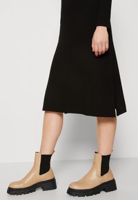 Beige nahkasaappaat nilkkakorkeudella mustilla elastisilla sivupaneeleilla, paksulla mustalla pohjalla ja sileällä pinnalla, yhdistettynä mustaan ribbimekkoon.