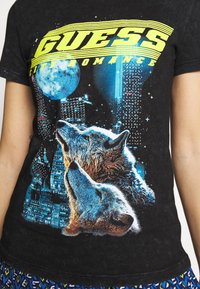 Dos lobos aullando a una luna azul con un fondo de paisaje urbano y texto amarillo "GUESS City Romance" en una camiseta negra.