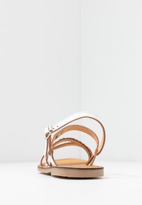 Witte leren sandaal met verstelbare enkelband, platte bruine zool en gevlochten details. Eenvoudig ontwerp met minimale hardwareaccenten.