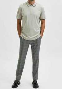 Polo gris clair avec col zippé, associé à un pantalon gris à carreaux et des chaussures noires. L'ensemble présente un design à la fois décontracté et structuré.