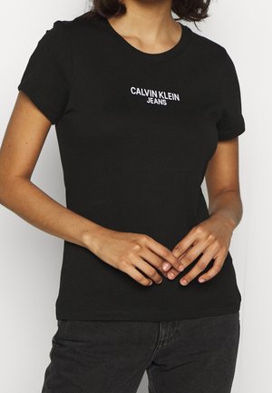 Persoon die een zwart Calvin Klein Jeans T-shirt met korte mouwen en een donkere broek draagt, handen zachtjes in de taille gevouwen.