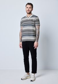 Pruhované polo tričko v béžové a černé barvě, krátké rukávy a límec. Nosené s černými kalhotami a světlými teniskami. Viditelné tetované ruce.