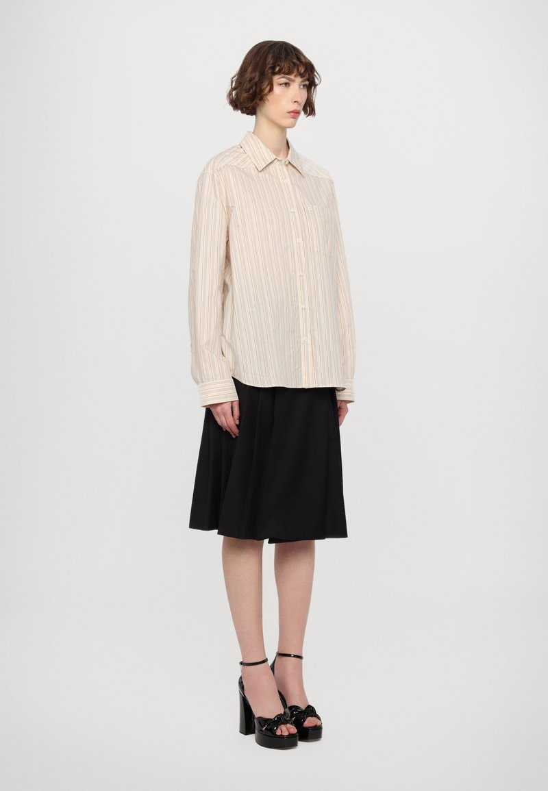 Blusa de rayas claras con cuello puntiagudo, cierre de botones y mangas largas; combinada con una falda plisada negra y sandalias de tacón negro.