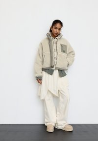 Fleece-baseret jakke i lysebeige med en grå panel, oversized pasform, lynlåslukning og ribkanter, lagdelt over en ternet skjorte og cremefarvede bukser.