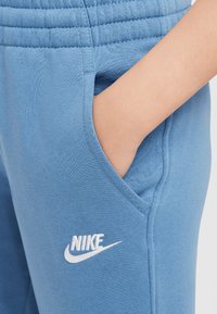Šviesiai mėlynos sportinės kelnės su tekstūruotu juosmeniu, turinčios šonines kišenes ir balta išsiuvinėta Nike logo kairėje pusėje.