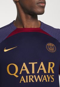 Camiseta deportiva de Nike en azul marino, con acentos en burdeos en los hombros, logo y texto dorado "QATAR AIRWAYS," tela texturizada en los hombros.