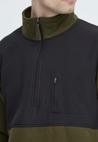 Pulóverová bunda s polovičným zipsom v čiernej a olivovej zelenej farbe. Značky fleece golier, zipsová náprsní kapsa a šité akcenty.