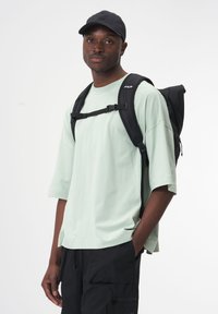 Schwarzer Rucksack mit Rolltop-Design und verstellbaren Trägern, getragen über einem mintgrünen Oversize-T-Shirt, kombiniert mit schwarzen Cargo-Hosen.