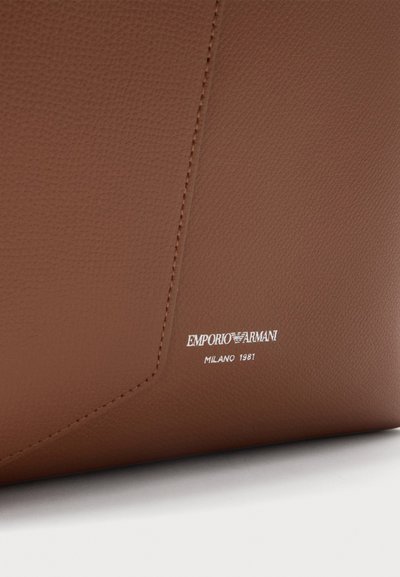 Καφέ δερμάτινο αντικείμενο Emporio Armani με λεπτή υφή, ορατά ράμματα και λευκό κείμενο λογότυπου που γράφει "EMPORIO ARMANI MILANO 1981" στον κάτω δεξιό μέρος.