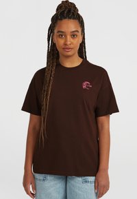 T-shirt en coton marron avec un col rond et des manches courtes, arborant un petit logo graphique rose sur la zone gauche de la poitrine.