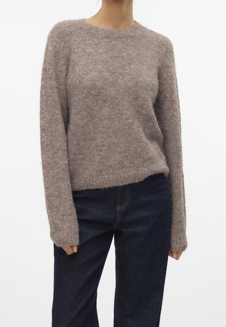 Femme portant un pull taupe à manches longues et duveteux, associé à un jean bleu foncé, debout devant un fond blanc uni.