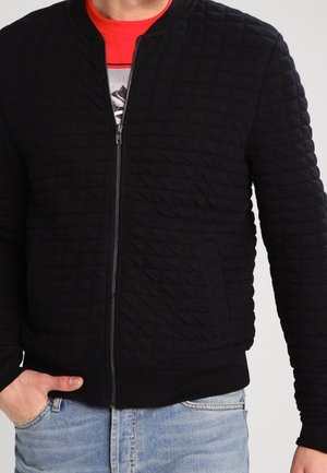 Uomo che indossa una giacca nera imbottita con cerniera sopra una maglietta rossa con stampa e jeans azzurri, con una mano in tasca, su sfondo semplice.