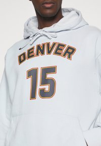 Hombre que lleva un sudadera ligera gris con "DENVER" y el número "15" impresos en azul marino contorneados en rojo y amarillo.
