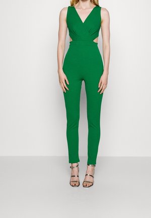 Jumpsuit vert, sans manches, avec un décolleté en V profond, des côtés découpés et des jambes ajustées. Accompagné de talons hauts argentés à lanières, texture lisse.