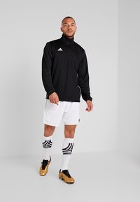 Svart zip-up treningsjakke, hvite shorts, svart og hvit stripete sokker, og gull støvler. Glatt stoff og sporty design.