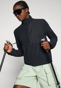 Schwarzer Fleece-Pullover mit Viertelreißverschluss und einer Reißverschlusstasche, getragen mit hellgrünen Cargo-Shorts. Hält Wanderstöcke und ist mit übergroßen Sonnenbrillen accessorized.