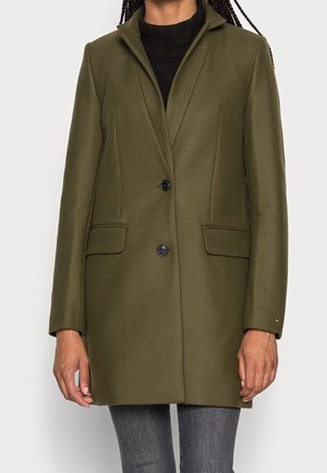 Frau trägt einen olivgrünen Wollmantel mit zwei schwarzen Knöpfen, Klappentaschen und einem kleinen Logo am linken Ärmel, kombiniert mit dunkelgrauen Hosen.