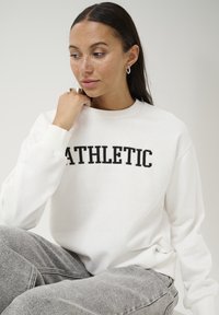 White sweatshirt with "ATHLETIC" in black bold font. Soft fabric, crew neck, and ribbed cuffs. Paired with gray sweatpants.