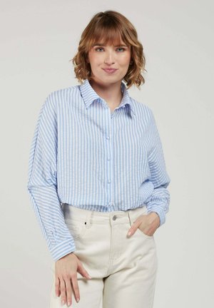 Femme aux cheveux courts et ondulés portant une chemise à rayures bleu clair et blanc boutonnée et un pantalon écru, posant avec une main dans la poche.