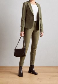 Marynarka w oliwkowej zieleni i dopasowane spodnie w tym samym kolorze, zestawione z czarną torebką i czarnymi botkami, na tle jasnoszarym.