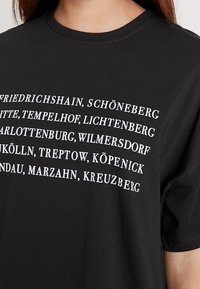 Camiseta de algodón negra con texto blanco que enumera varios distritos de Berlín en una fuente sans-serif negrita. Mangas cortas y cuello redondo.