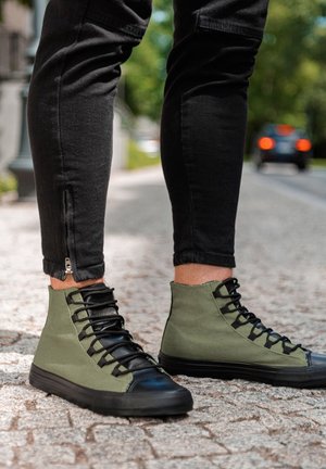 Baskets montantes en toile vert olive avec des accents en cuir noir. Dotées de lacets noirs, d'une semelle en caoutchouc texturée et de détails zippés sur le pantalon.