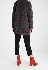 Grijze nepbontjas met een gemêleerd, gewatteerd patroon; gecombineerd met zwarte broek en rode enkellaarsjes, wat een hedendaagse silhouet toont.