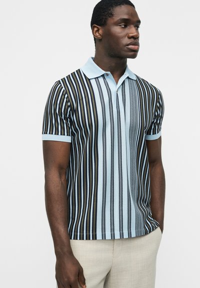 Νεαρός άνδρας που φοράει κοντομάνικο πόλο με κάθετες μαύρες, καφέ και ανοιχτές μπλε ρίγες, συνδυασμένο με μπεζ παντελόνι, στέκεται μπροστά σε λευκό φόντο.