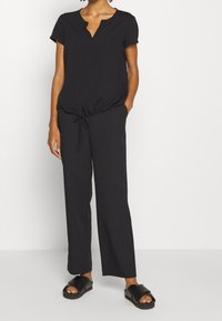 Haut noir à manches courtes avec col en V et une poche, associé à un pantalon noir ample et des sandales plates noires. Tissu doux et lisse.