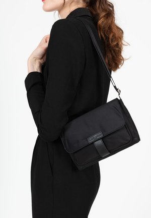 Borsa a spalla nera realizzata in tessuto liscio con un accento in pelle. Presenta una chiusura a patta e una tracolla regolabile, mantenendo una forma compatta e rettangolare.