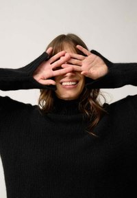 Czarny, dzianinowy sweter z golfem i długimi rękawami z otworami na kciuki. Ręce modelki zasłaniają jej oczy, co pokazuje odprężony wyraz twarzy.