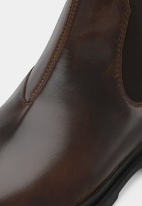 Brown leather ankle boot with a smooth finish, elastic side panel, and a rounded toe. Features contrast stitching and a rubber sole.