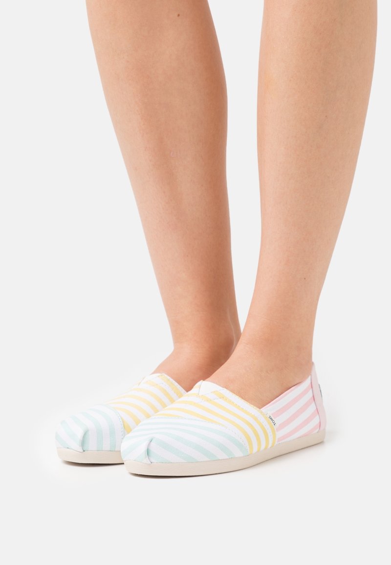 Pieds chaussés de chaussures sans lacets avec des motifs à rayures pastel en bleu, jaune et rose sur une semelle claire, présentés sur un fond uni.