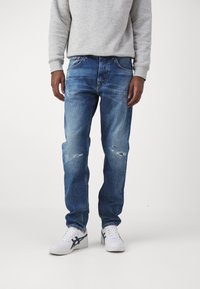 Blå denimjeans med en afslappet pasform, der har slidte detaljer, let falmning og fem-lommer design, kombineret med hvide sneakers.