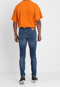 Jeans de mezclilla azul con corte slim, que cuentan con bolsillos traseros y un parche de cuero en la cintura, combinados con una camiseta de gran tamaño color naranja.